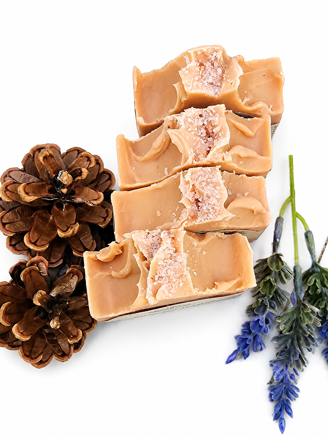 Bar of soap with a natural texture, surrounded by pine cones and lavender on a white background. Topped with pink sea salt