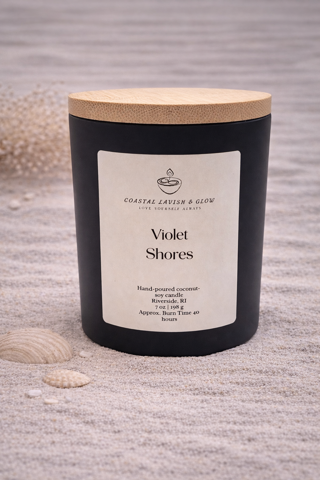 Violet Shores black glass candle with a wooden lid and label on a textured surface with shells.  Lavender and sage scent.