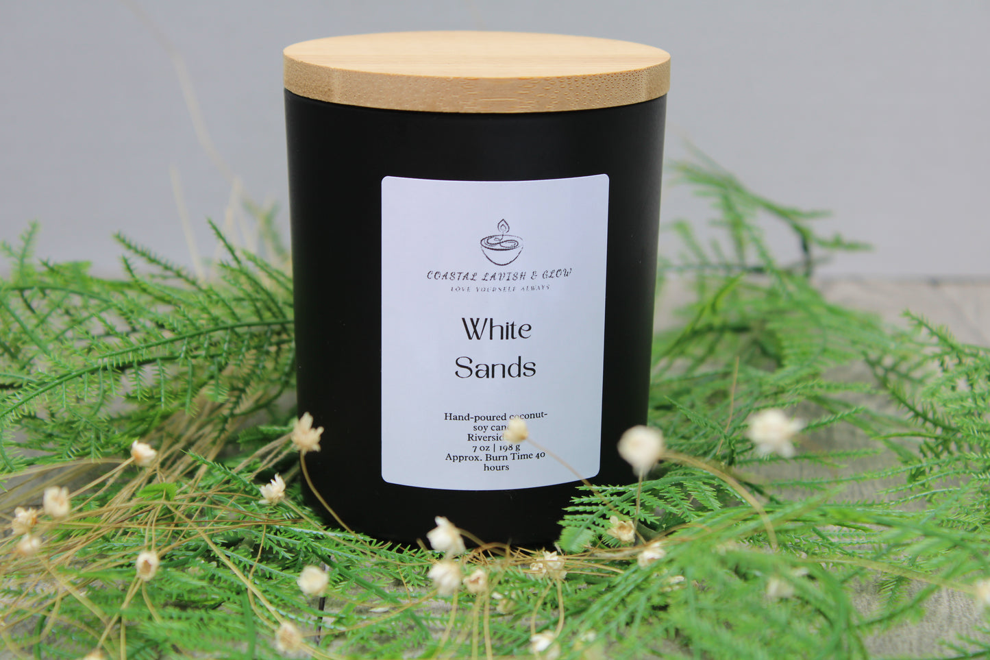 Black candle labeled 'White Sands' with a wooden lid on green foliage.